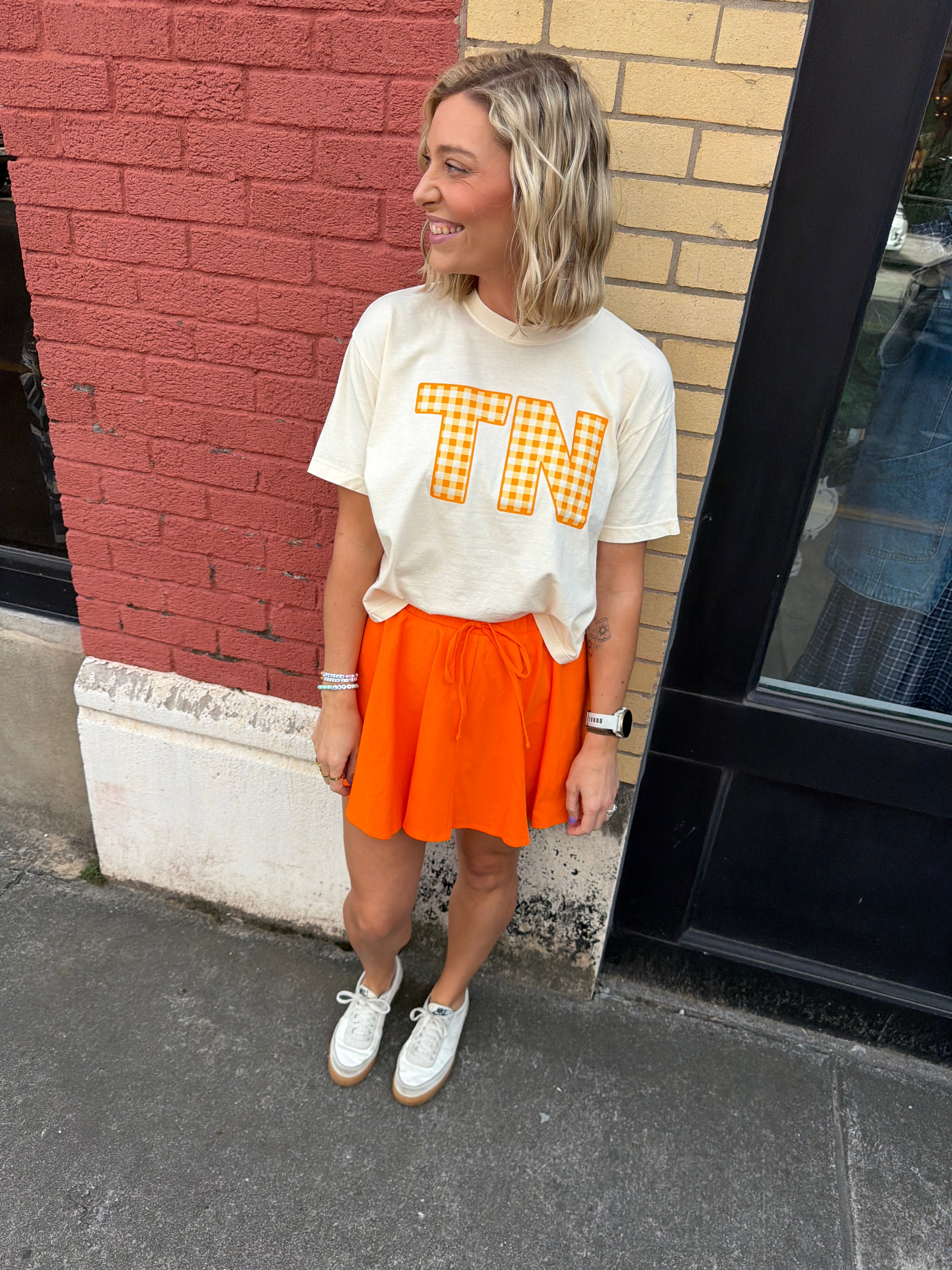 Person wearing a short sleeve cream t-shirt with  a checkered 'TN' printed on it and an orange skirt standing on a sidewalk.
