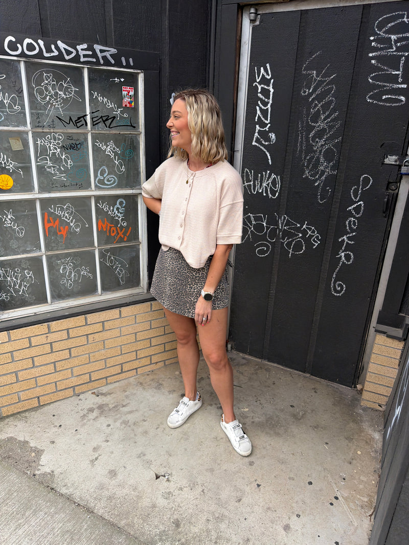 woman standing on sidewalk with a leopard skort on with a tan button up short sleeve sweater.